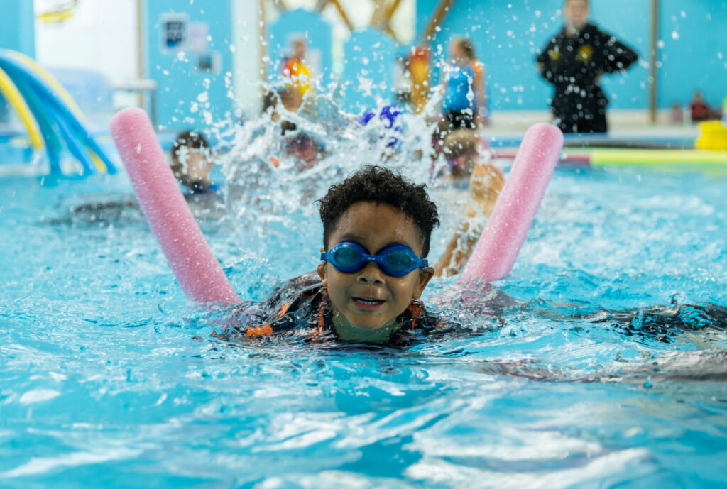 Photo of young boy swimming in pool with noodle as swim teacher guides with instruction